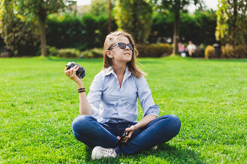 Girl listening music to portable speaker while sitting outdoors - Millennial having fun in the park with small wireless entrainment device - Cute female using technology in nature