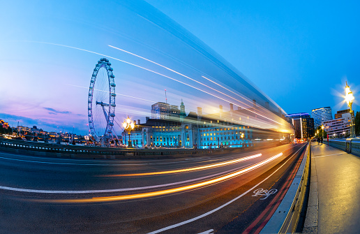 London, England, United Kingdom - July 16, 2019: Long exposure photography of Double decker red bus in blurred motion on Westminster Abbey bridge against the Eye Wheel
