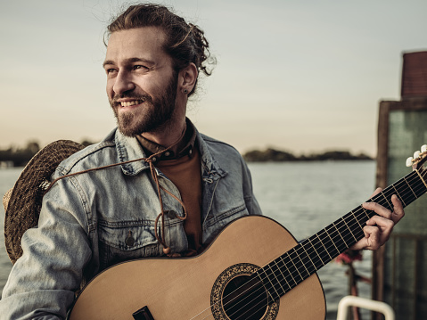 Portrait of the young man with guitar looking over river water. End of the summer weekend.