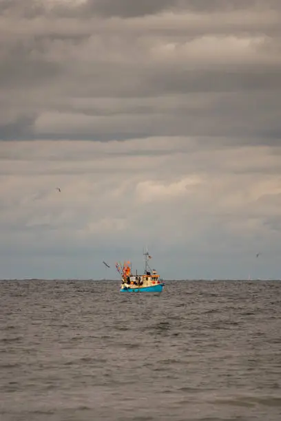 Photo of far on the sea floats a fishing boat, which is surrounded by many seagulls and the sky is full of clouds