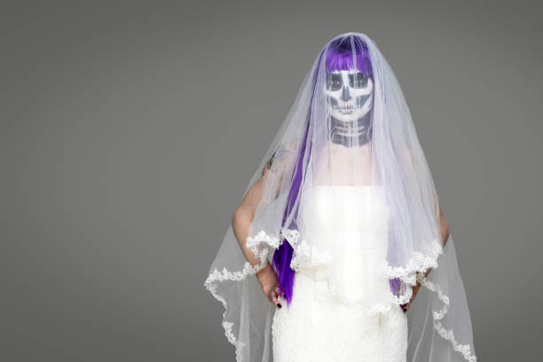 porträt der frau schaut auf die kamera mit erschreckenden halloween skelett make-up und lila perücke brautschleier, hochzeitskleid über grauem hintergrund. schwarze hochzeit - day of the dead mexico bride human skeleton stock-fotos und bilder