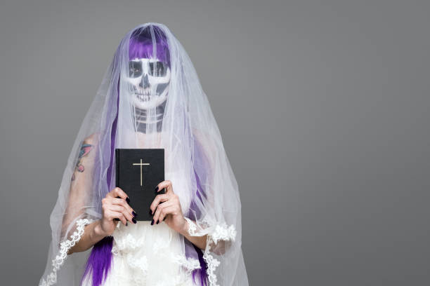 porträt der frau schaut auf die kamera mit erschreckenden halloween skelett make-up und lila perücke brautschleier, brautkleid, hält die heilige bibel über grauen hintergrund. schwarze hochzeit. kopierraum - day of the dead mexico bride human skeleton stock-fotos und bilder