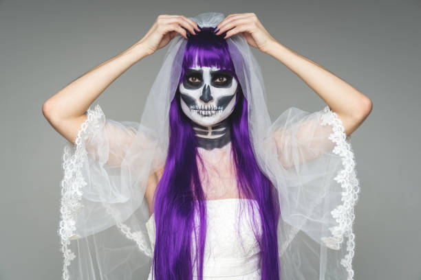 Portrait of woman looks at the camera with terrifying halloween skeleton makeup and purple wig corrects bridal veil, wedding dress over gray background. Black wedding. stock photo