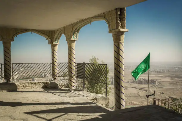 Desert city view from the mosque on top of the mountain in Qom city, Iran. Columns, fence, arcade and green flag on a background of blue sky.