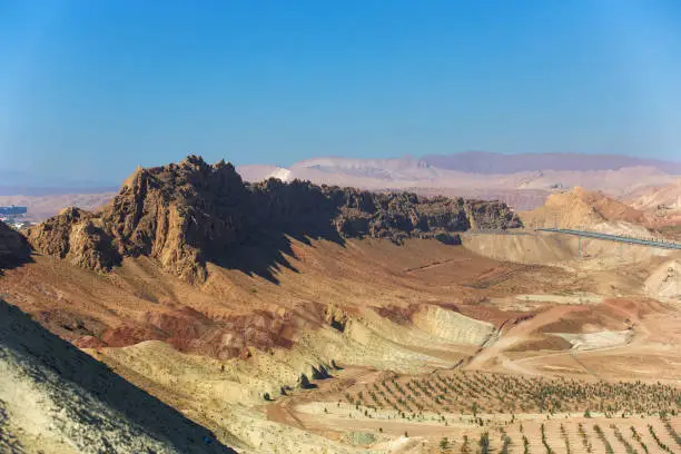 Desert landscape with red and yellow cliffs, rows of plants and mountains in the background. View of the outskirts of Qom in Iran