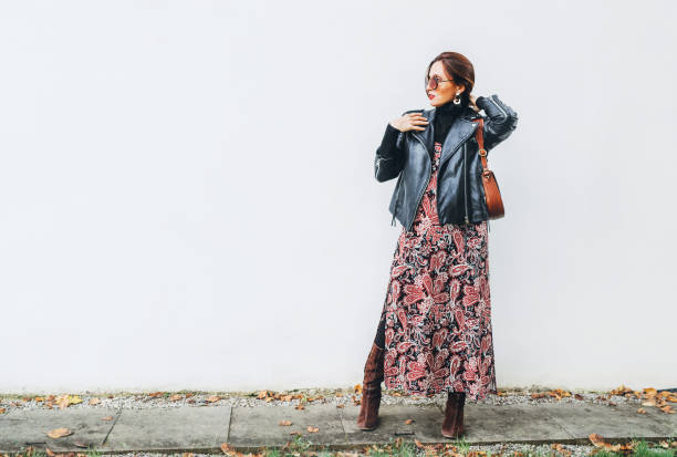 smiling female dressed boho fashion style colorful long dress with black leather biker jacket with brown leather flap bag posing on the white wall background. - clothing fashion model old fashioned women imagens e fotografias de stock