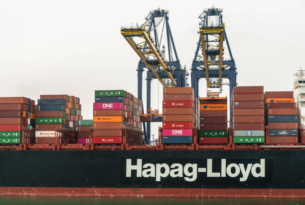 Laem Chabang, Thailand - March 16, 2019: Container port and terminal and mid section of Hapag Lloyd ship under gray sky. Two yellow cranes loading and unloading it. Colored container stacks on ship.