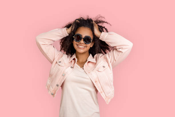 kür. afrikanische mädchen in ausgefallenen sonnenbrille jeansjacke stehen isoliert auf rosa halten haare fröhlich - afro amerikanischer herkunft fotos stock-fotos und bilder