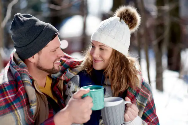 couple of man and woman having a romantic picnic date in snow during the winter season, and cuddling under a blanket and sharing a cup of cocoa to keep warm