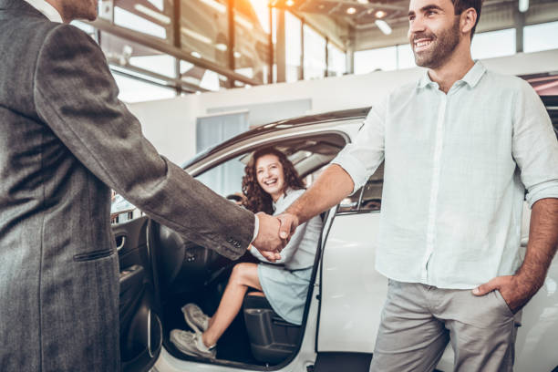 close up view of handshake of salesmen with the man in the showroom on the background of a woman in a new car close up view of handshake of salesmen with the man in the showroom on the background of a woman in a new car. saloon car stock pictures, royalty-free photos & images