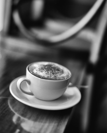 Analogue black and white image of cup of cappuccino sitting on a table in a coffee shop, with shallow depth of field