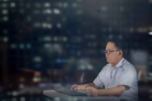 A calmly working student inside an office shot during night time in the city. foto de vista lateral de un joven estudiante que trabaja en su computadora portátil en su escritorio por la noche dentro de su sala de oficina. hombre con uniforme de polo con gafas solo en una habitación. trabajando en el turno de noche.