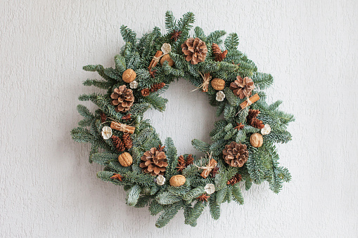 Christmas wreath made of natural fir branches  hanging on a white wall.  Wreath with natural ornaments: bumps, walnuts, cinnamon, cones. New year and winter holidays. Christmas decor