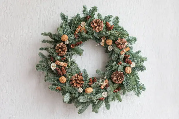 Photo of Christmas wreath made of natural fir branches  hanging on a white wall.  Wreath with natural ornaments: bumps, walnuts, cinnamon, cones. New year and winter holidays. Christmas decor