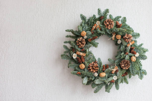 Christmas wreath made of natural fir branches  hanging on a white wall.  Wreath with natural ornaments: bumps, walnuts, cinnamon, cones. New year and winter holidays. Christmas decor. Copy space stock photo