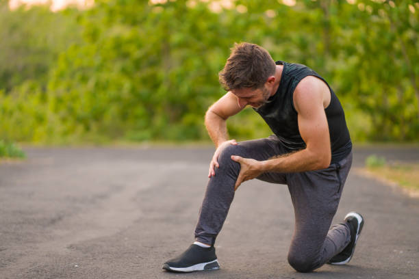 outdoors portrait of young attractive sport man in pain touching his knee suffering ligaments accident or some injury during running workout at beautiful country road in health care concept stock photo