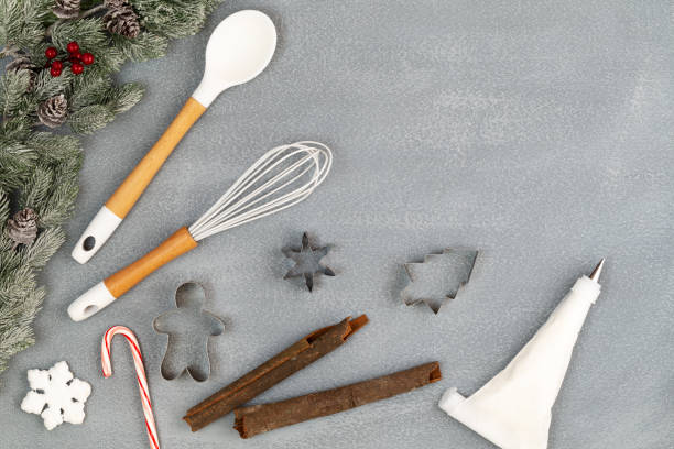 Utensils and spice for Christmas cooking or baking with spoon, whisk, icing piping bag, cookie cutters, cinnamon sticks, candy cane and snow flake with snowy fir branches over stone like background with copy space. Flat lay, top view. stock photo