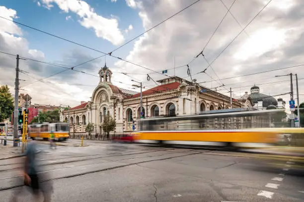 Day time long exposure cityscape in downtown Sofia, capital city of Bulgaria, Eastern Europe. Including the main building of the Central Market Hall (covered market) in Sofia since 1911 and is today an important trade centre in the city. Photographed at the cross roads with blurred lights from the road traffic and passing yellow trams on the Marie Louise Boulevard. Shot on Canon full frame EOS R full frame system with tripod. Image is great for background with extra copy space for your message.