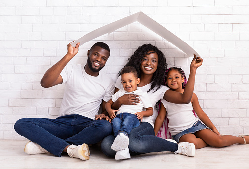 Family Housing Concept. Young African American Parents With Two Children Sitting Under Cartboard Roof Dreaming Of New Home, Copy Space