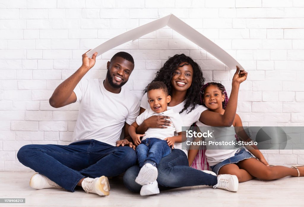 Beautiful Black Family Holding Cardboard Roof Dreaming Of New Home Family Housing Concept. Young African American Parents With Two Children Sitting Under Cartboard Roof Dreaming Of New Home, Copy Space Family Stock Photo