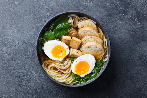 Asian noodle soup, ramen with chicken, tofu, vegetables and egg in black bowl. Top view. Copy space. Slate background