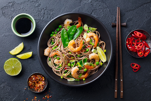 Stir fry noodles with vegetables and shrimps in black bowl. Slate background. Top view.