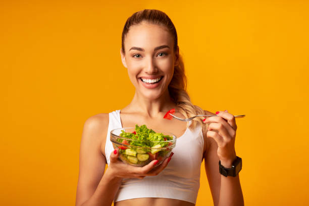 fitness chica comiendo ensalada de verduras de pie sobre fondo amarillo - healthy eating sport exercising women fotografías e imágenes de stock