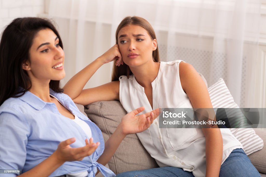 Envious Girl Listening To Friend Sitting On Couch At Home Envy. Jealous Girl Listening To Her Friend Talking About Her Great Life Sitting On Couch At Home. Selective Focus Friendship Stock Photo