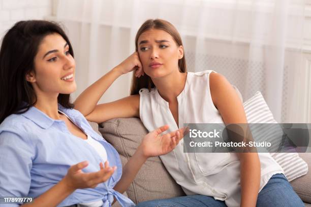 Envious Girl Listening To Friend Sitting On Couch At Home Stock Photo - Download Image Now