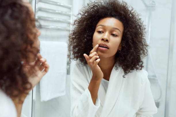 Lips skin care. Woman applying lip balm in bathroom portrait Lips skin care. Woman applying lip balm looking in mirror at bathroom. Portrait of beautiful african girl model with beauty face and natural makeup applying lip product with finger human lips stock pictures, royalty-free photos & images