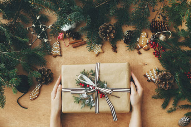 istock Merry Christmas flat lay. Hands holding stylish rustic christmas gift and pine branches, cones, gingerbread cookies,thread, cinnamon, anise on rural wooden table. Seasons greeting 1178927005