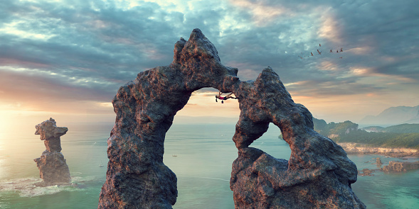 A wide angle panoramic image of a female extreme free climbers upside down, traversing the underside of a huge natural rock arch. The climber is wearing a top, shorts, climbing shoes and chalk bag . The location is fictional, by the sea as the dawn sun rises.