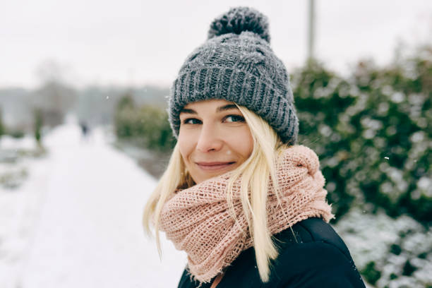Close-up portrait of blonde beautiful young woman smiling wearing winter jacket, gray knitted hat and pink scarf, posing outdoors in the park on snowing winter day. Christmas mood. stock photo