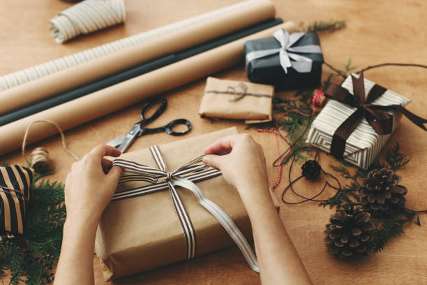 istock Hands wrapping stylish christmas gift box in craft paper and scissors, rustic  presents, thread, pine branches and cones on wooden table. Wrapping christmas gifts 1178934937