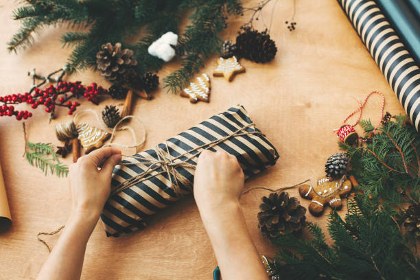 istock Hands wrapping christmas gift in stylish striped paper and pine branches, cones, gingerbread cookies, thread, cinnamon on rural wooden table. Wrapping christmas present 1178935202
