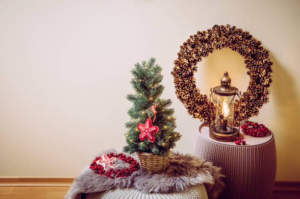 istock Small portable tabletop decorated Christmas tree on table, party lights illuminated, red felt fabric ornament. Light bulb lantern with pine cone wreath on background. Copy space for Christmas card. 1178942482