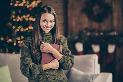 Close up photo of positive cheerful brown hair girl in knitted jumper hold fiction, book enjoy her jolly christmas celebration feel content in house full of newyear x-mas decorations lights indoors