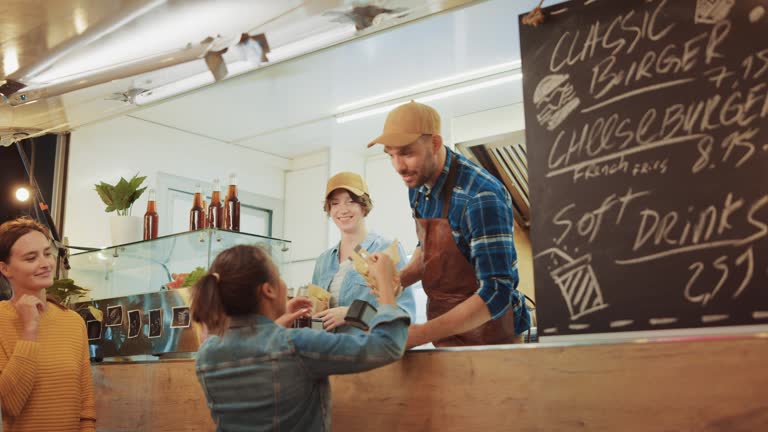 Food Truck Employee Hands Out a Freshly Made Burger to a Happy Young Female. Young Lady is Paying for Food with Contactless Credit Card. Street Food Truck Selling Burgers in a Modern Hip Neighbourhood