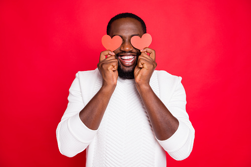 Portrait of positive cheerful expression funky dark skin man hold small paper card heart close eyes want surprise girlfriend date 14-february, lover wear white clothes isolated red color background