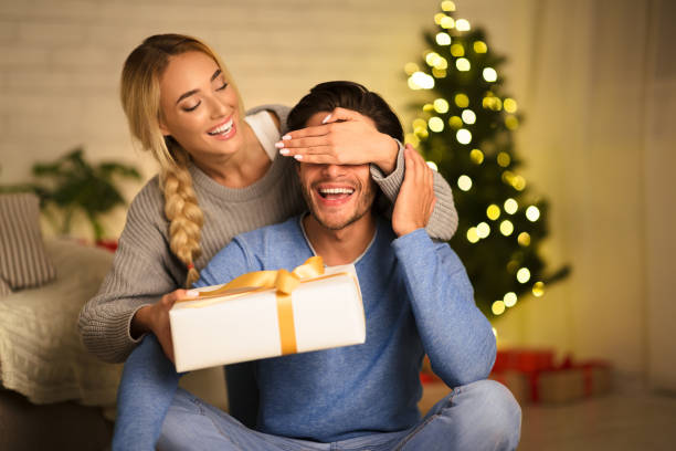 Christmas gift. Happy woman making surprise to husband Christmas gift. Happy woman making surprise to husband, closing his eyes in front of christmas tree unwrapping stock pictures, royalty-free photos & images