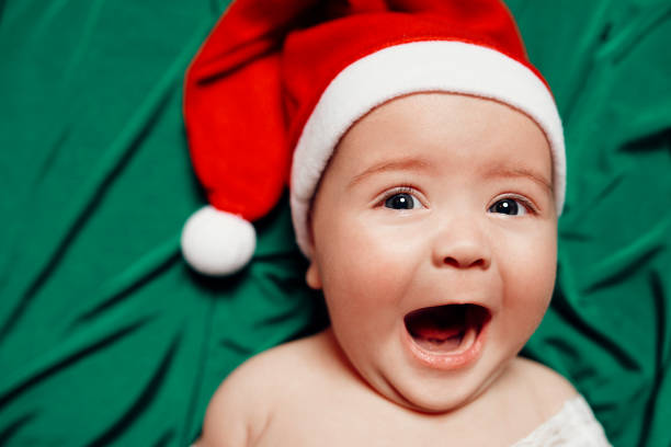 Little baby girl lying on her back on green bed, wearing Christmas hat and looking at the camera screaming