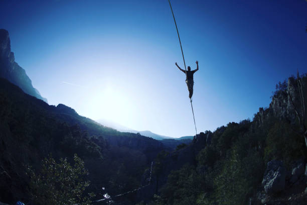 a man is walking along a stretched sling. highline in the mountains. man catches balance. performance of a tightrope walker in nature. highliner on the background of the mountains. - form fitted imagens e fotografias de stock