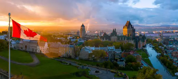 Aerial panorama view of Old Quebec City. To left side of frame is a Canadian flag in front of a beautiful golden sunset. To the right of the frame is Old Quebec, including the Chateau Frontenac and Defferin Terrace.