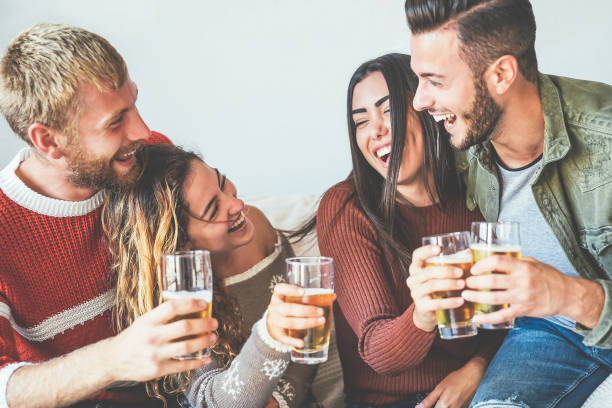 group of happy friends cheering with beer at home - millennial young people having fun drinking and laughing together sitting on sofa - friendship, entertainment and youth lifestyle holidays - life events laughing women latin american and hispanic ethnicity imagens e fotografias de stock