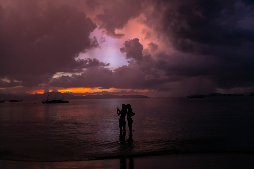 At dusk, two Filipino women stand in shallow water at the beach while looking at a smartphone in Port Barton, Palawan, Philippines