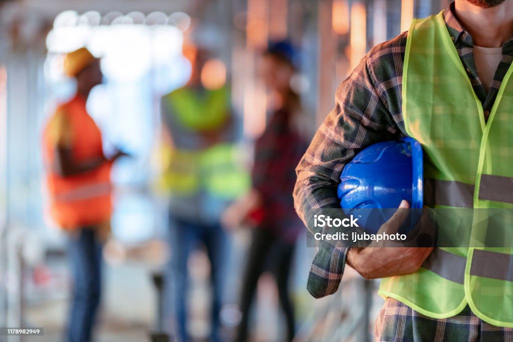 Man holding blue helmet close up Man holding blue helmet close up. Construction man worker with office and people in background. Close up of a construction worker's hand holding working helmet. Occupation Stock Photo
