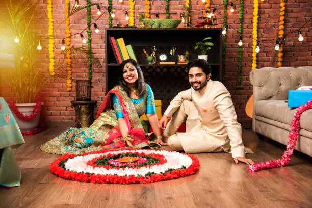 Photo of Indian couple making flower Rangoli on Diwali or Onam Festival, taking selfie or holding sweets