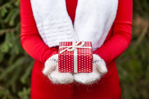 Girl hands in white knitted mittens holding a small handmade red Christmas giftbox with bow. Concept of Xmas or Birthday surprise.
