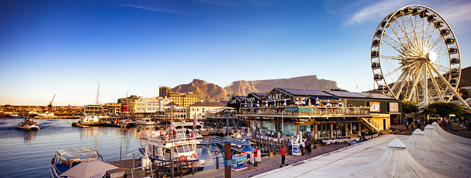 Cape town panoramic view of Victoria and Albert waterfront with table mountain in the background. In the foreground, the cape union mart, the Ferris wheel to the right and docked boats. A few people are visible enjoying the premises.