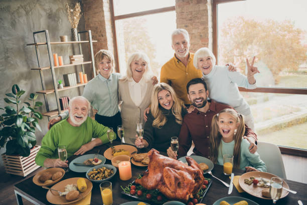 foto da família grande que está abraçando o feriado da tabela da festa da mesa peru roasted que faz o retrato parentes multi-generation que levanta a mostra dos vidros de vinho v-assine dentro a sala - thanksgiving dinner party feast day turkey - fotografias e filmes do acervo